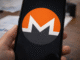 A smartphone held in hand displays the Monero cryptocurrency logo on its screen, with financial documents blurred in the background.